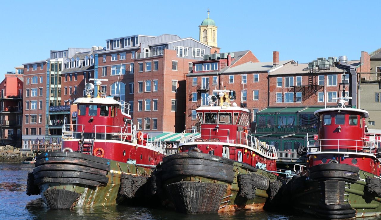Three red tugboats docked at the waterfront in Portsmouth, New Hampshire, with historic brick buildings and a clear blue sky in the background.