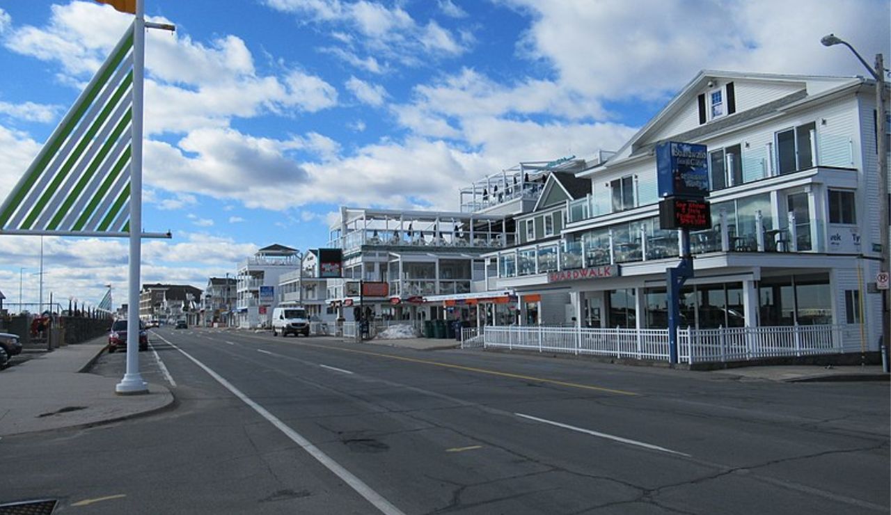 A quiet street along the Hampton Beach boardwalk in New Hampshire, lined with white beachfront buildings and restaurants under a partly cloudy blue sky.
