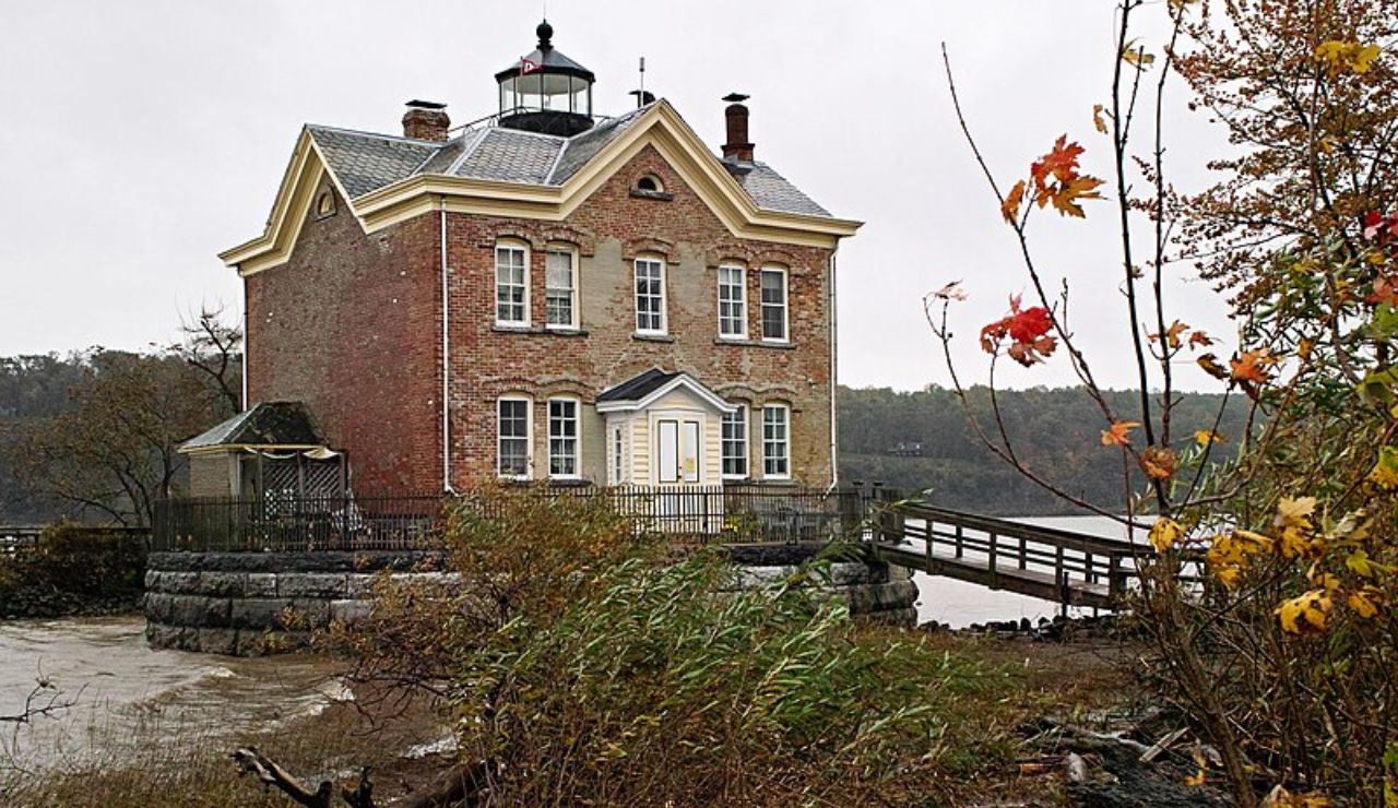 Historic brick lighthouse with a small tower, surrounded by autumn trees and overlooking the Hudson River in New York.