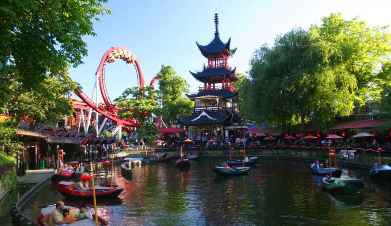 Amusement park with paddle boats on a scenic canal, a red roller coaster, and a pagoda-style building surrounded by trees.
