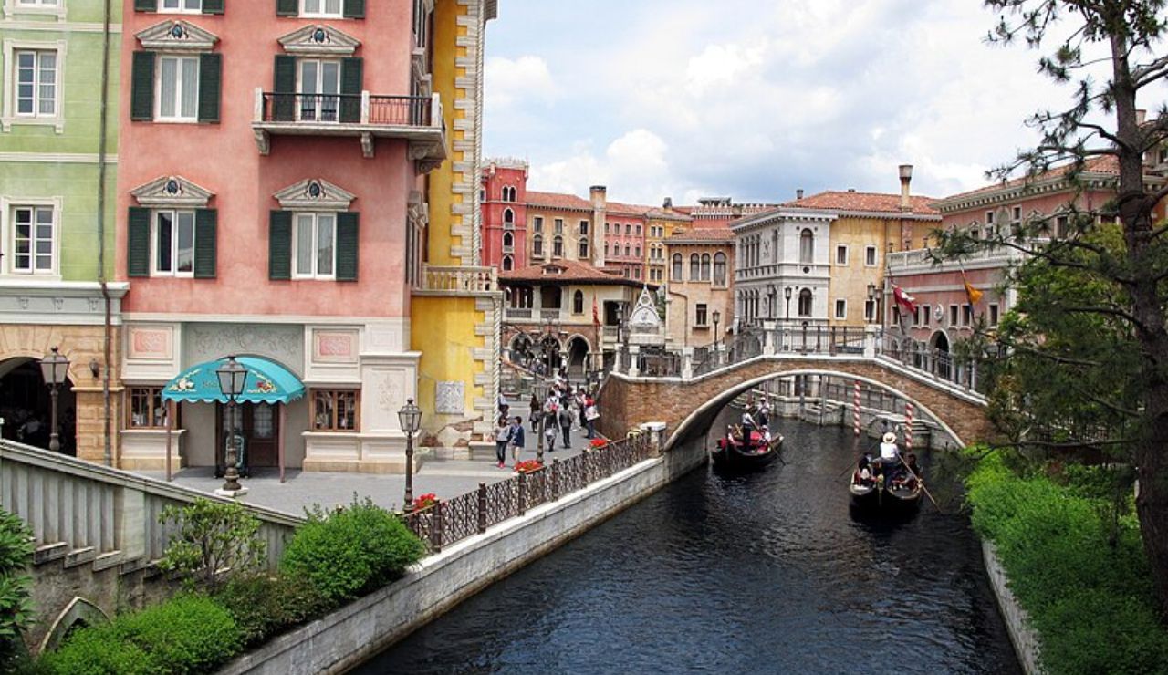 Colorful European-style buildings and gondolas on a canal with a stone bridge in a theme park setting.