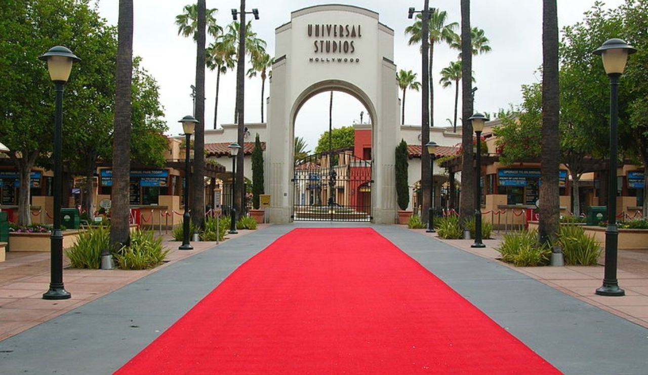 Red carpet leads to the iconic archway entrance of Universal Studios Hollywood, surrounded by palm trees and vintage-style lampposts.