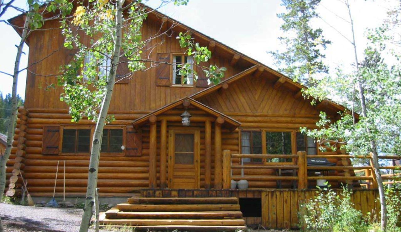 A large wooden log cabin with a sloped roof and front porch, surrounded by young trees and forest greenery.