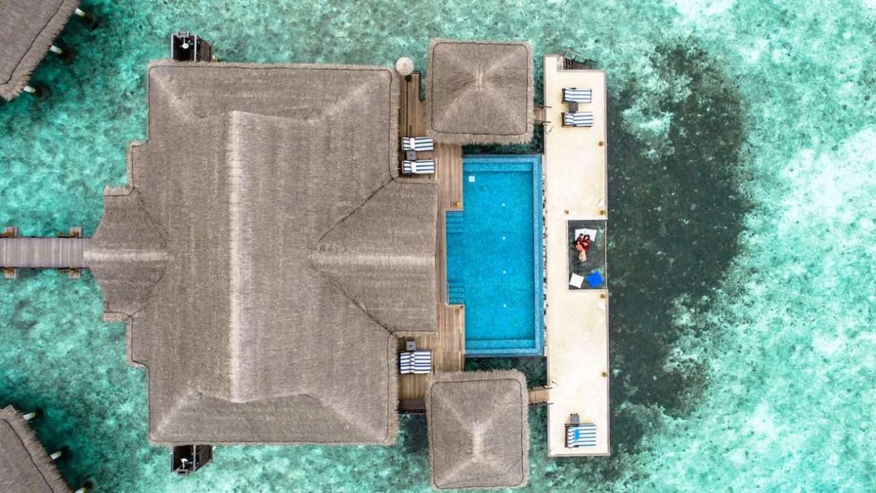 Aerial view of a luxury overwater villa with a private pool and a couple relaxing by the ocean