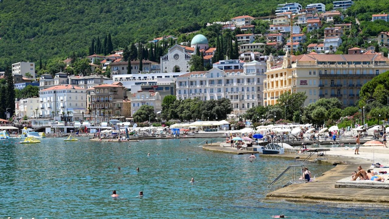 People swim and relax along a sunny seaside promenade lined with historic buildings and hillside homes