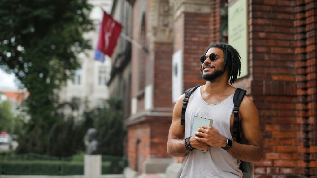 Smiling man with a backpack and notebook standing by a brick building.