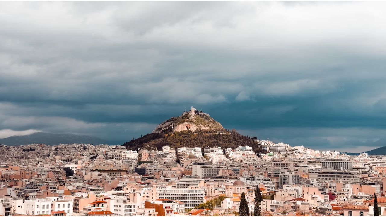 Cityscape of Athens with Mount Lycabettus rising in the center under a dramatic cloudy sky.