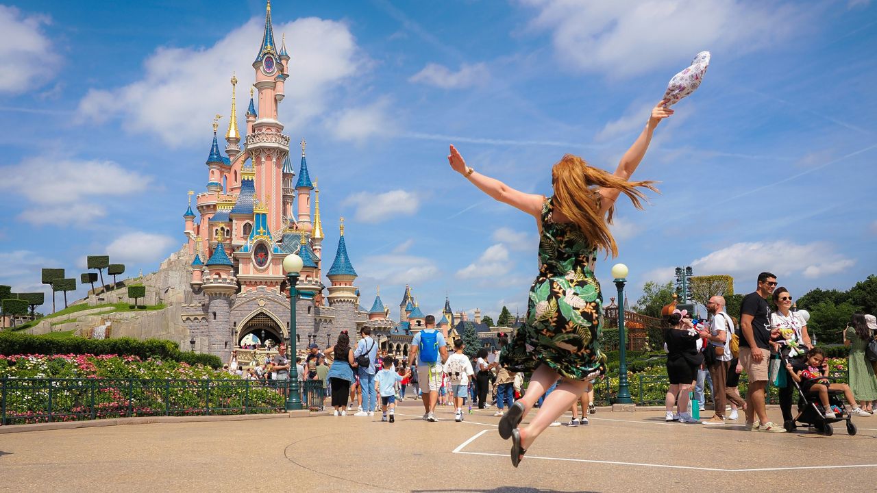 Woman joyfully jumping in front of a fairytale castle at a theme park, surrounded by tourists.