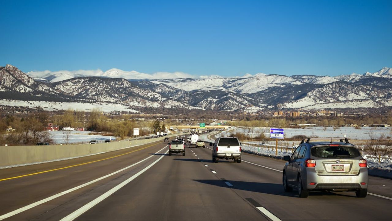 Cars driving on a highway with snow-covered mountains in the background under a clear blue sky.