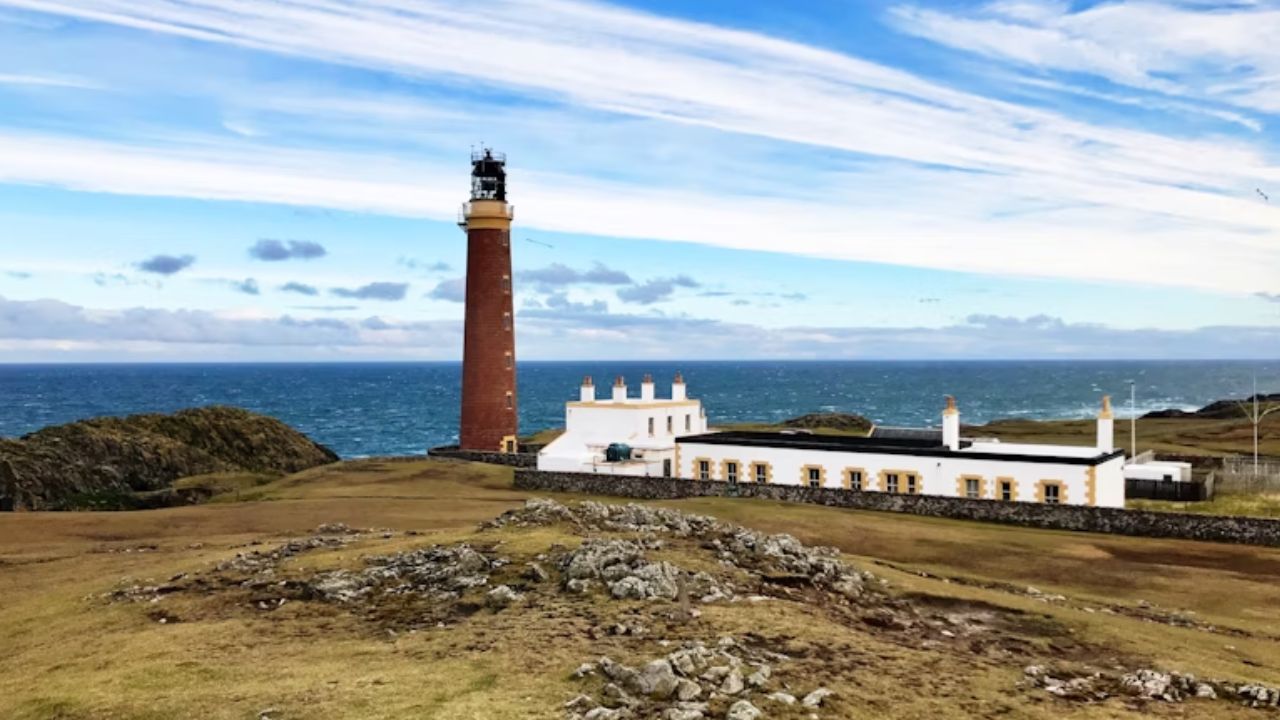 A tall red-brick lighthouse beside a long white keeper's house overlooking the rocky coast and open sea under a partly cloudy sky.