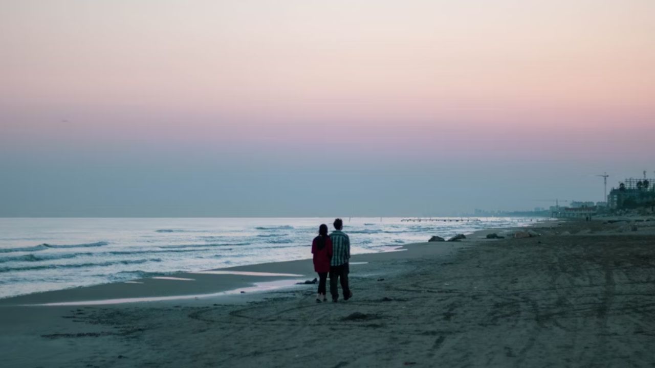 Couple walking along a quiet beach at sunset with soft pastel skies.