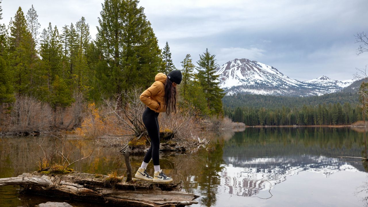Woman standing on a log by a calm lake, surrounded by pine trees and snow-capped mountains in the background.