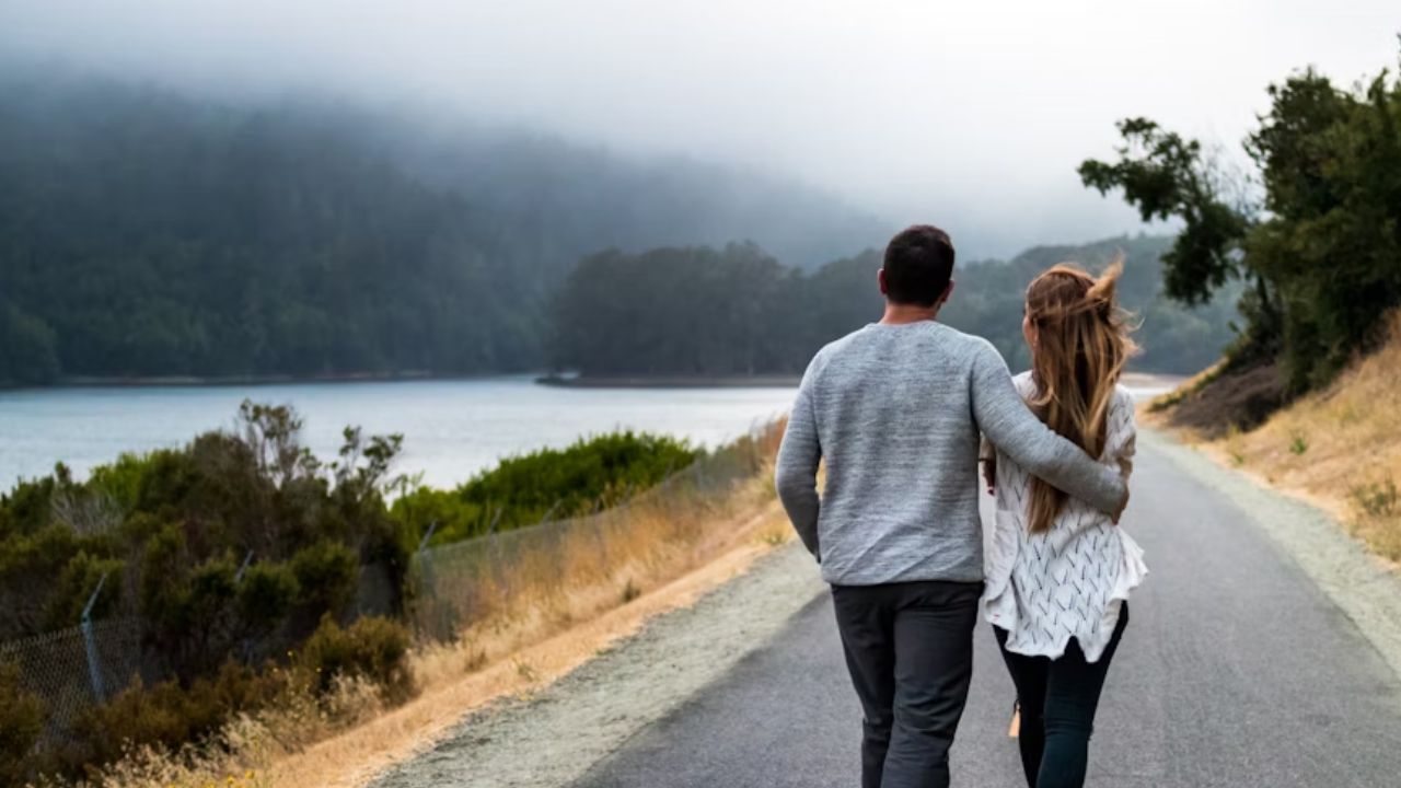 A couple walks arm-in-arm down a peaceful road beside a lake, surrounded by trees and hills under a misty sky.