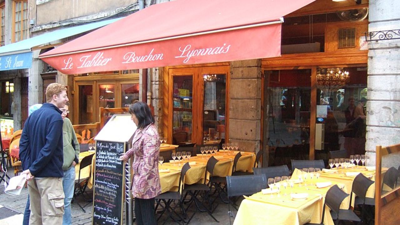 People stand outside a traditional French restaurant, "Le Tablier Bouchon Lyonnais," with a red awning and outdoor tables covered in yellow tablecloths.