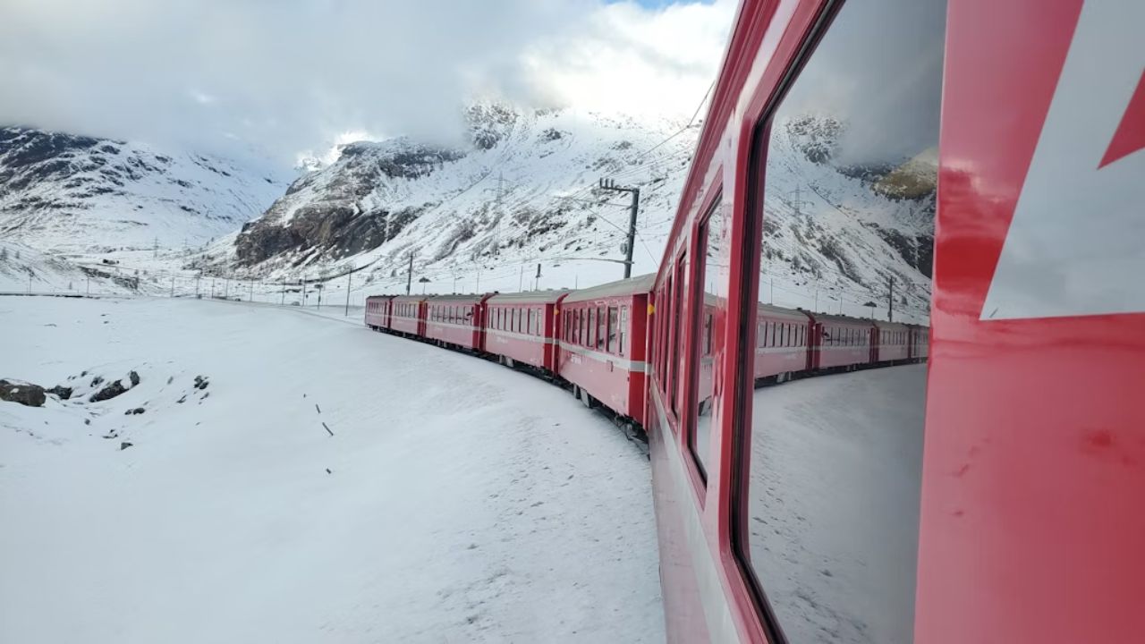 A red train winding through the snowy Swiss mountains, with its reflection visible in the window.