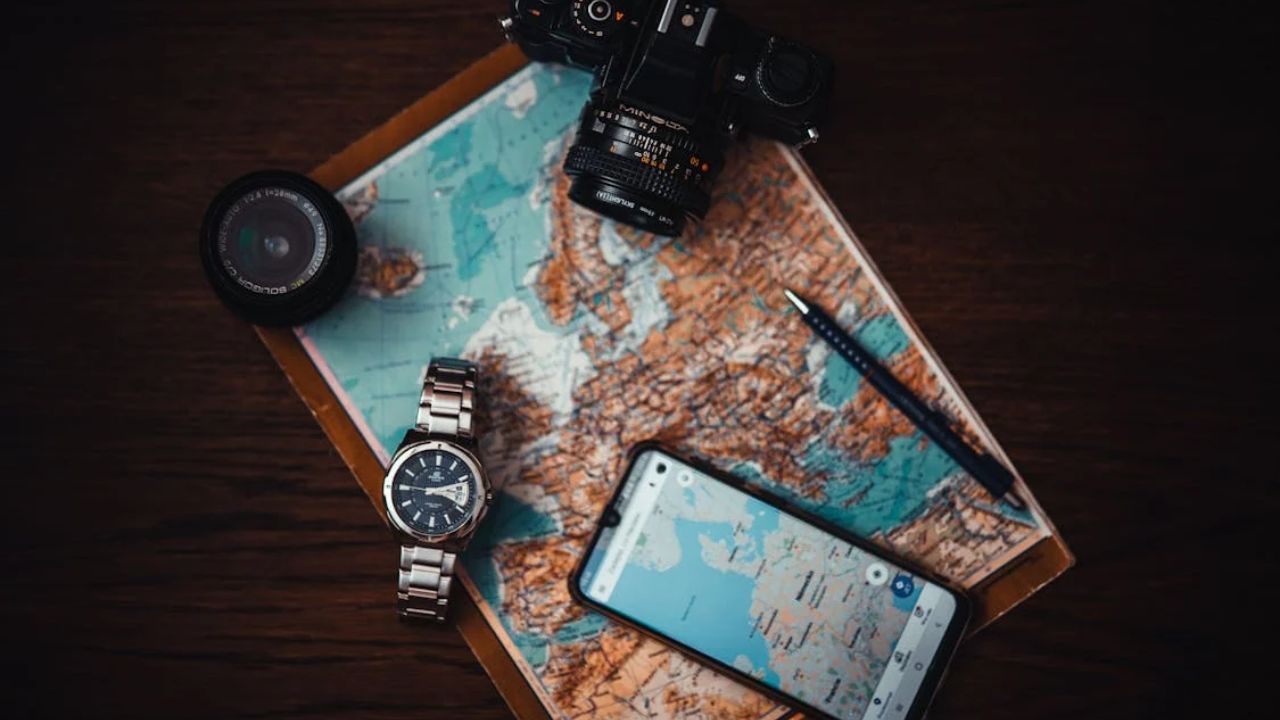 Flat lay of travel essentials including a map, smartphone with navigation app, camera, lens, watch, and pen on a wooden table.