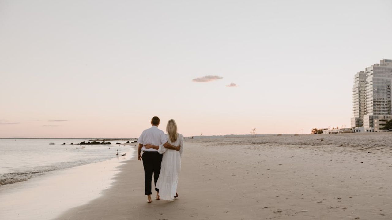 Couple walking hand-in-hand along a sandy beach at sunset with city buildings in the background.