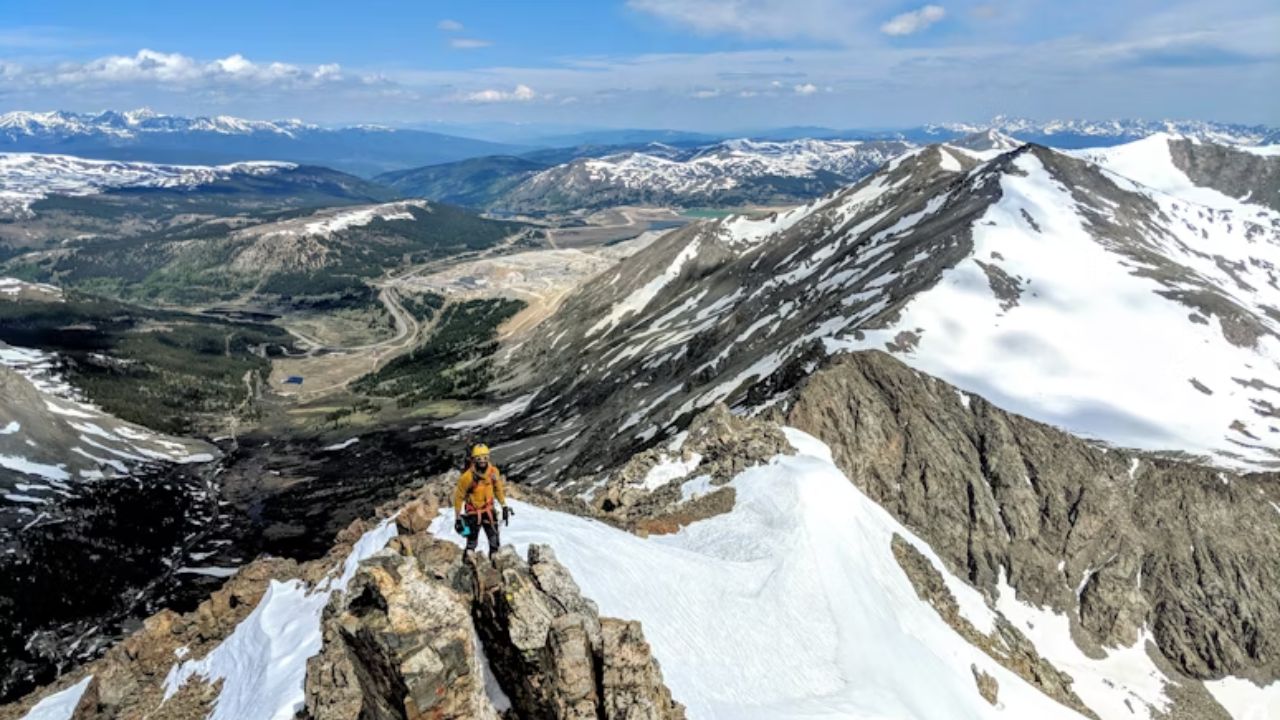 A climber in yellow gear standing on a narrow, snowy mountain ridge with panoramic views of surrounding peaks and valleys.