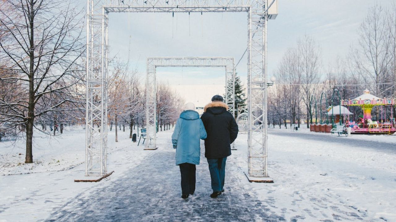 Couple walking together through a snowy park decorated with metal arches and winter trees.
