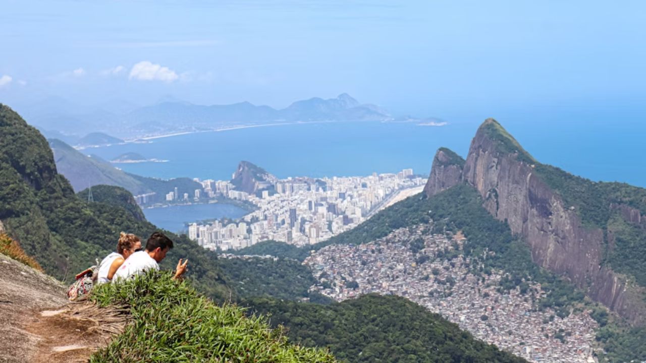 A couple sitting on a rocky ledge, enjoying the stunning view of Rio de Janeiro, with the city, coastline, and mountains in the background.