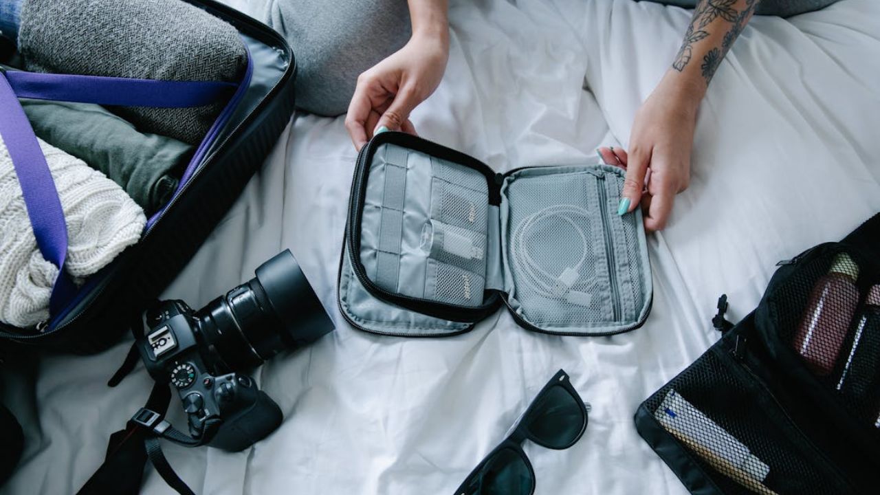 Person organizing travel essentials on a bed, including cables, toiletries, camera, and packed suitcase