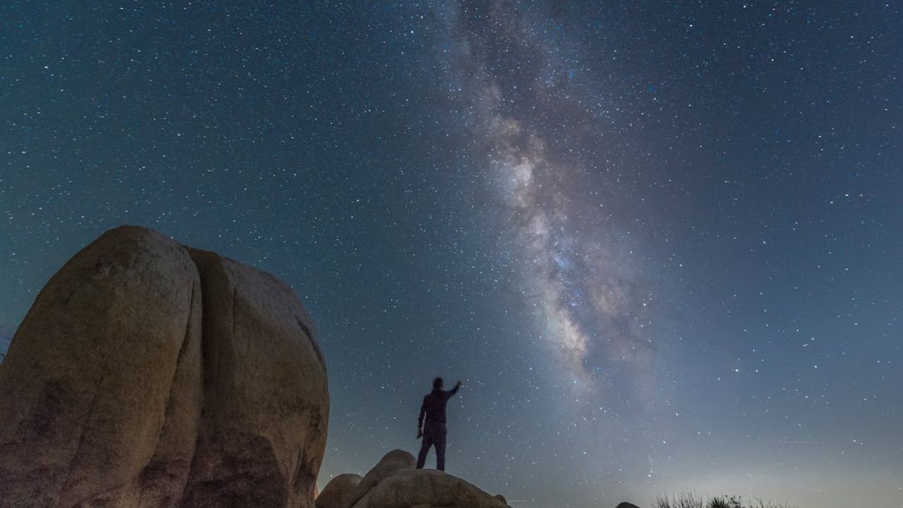 A person standing on rocks under a starry night sky, pointing toward the bright band of the Milky Way galaxy.