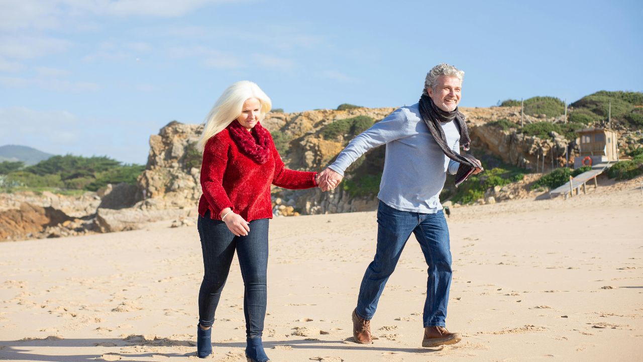 Older couple walking hand in hand on a sandy beach with rocky cliffs in the background.