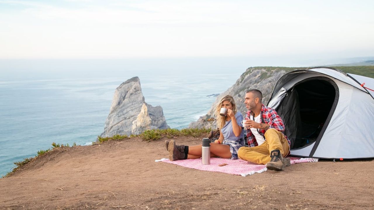 Couple camping on a cliffside with ocean view, sitting on a blanket near their tent and enjoying drinks.