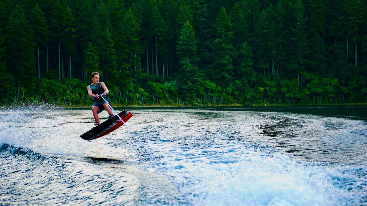 Person wakeboarding mid-air on a forested lake, creating splashes and wake trails.