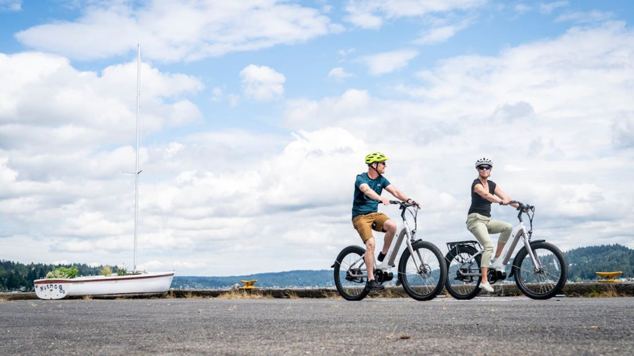 Man and woman ride electric bikes near a dock with a sailboat in the background.