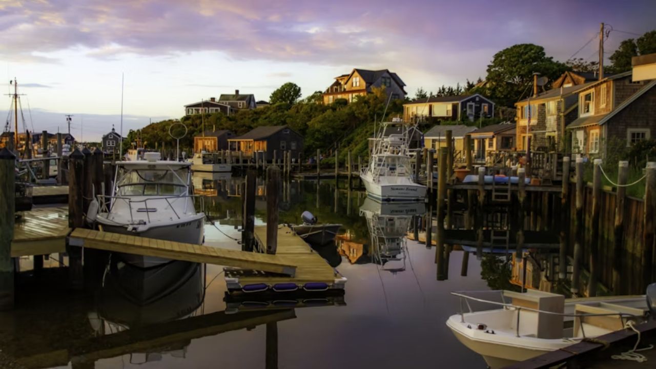 A peaceful harbor at sunset in Menemsha, Martha’s Vineyard, with fishing boats docked along wooden piers and coastal cottages reflecting on the still water.