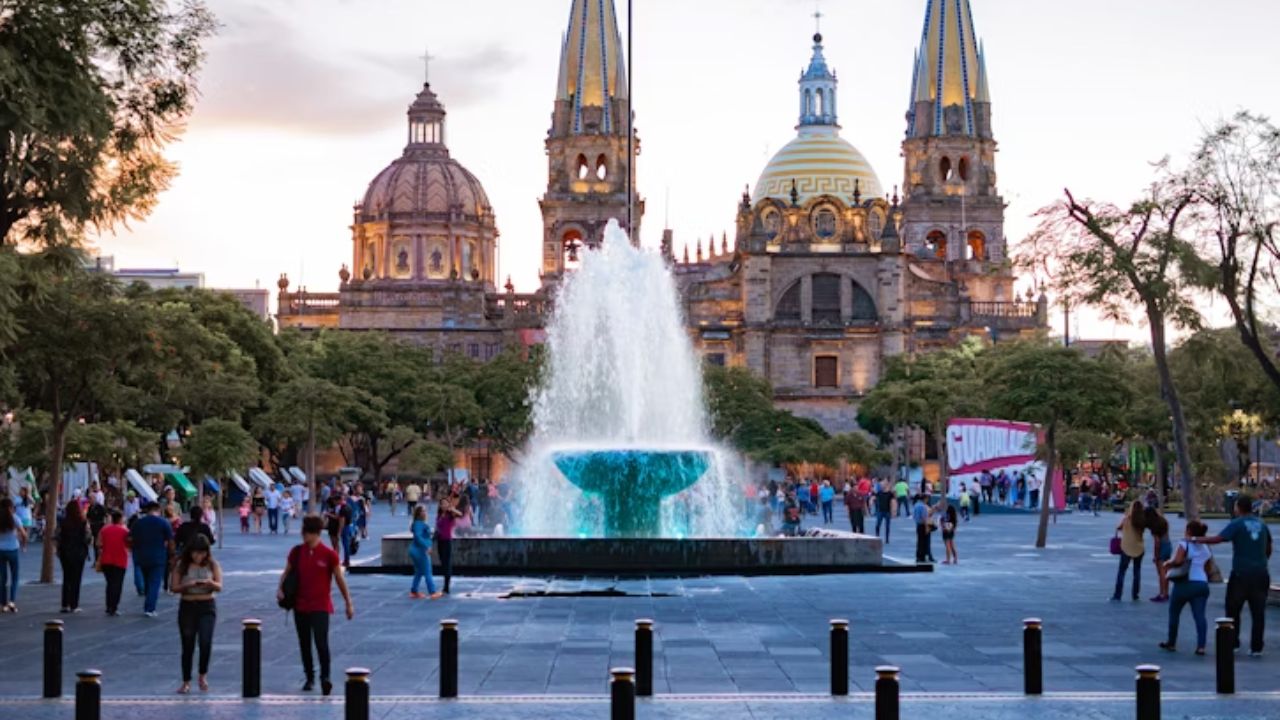 People walking near a large fountain with the Guadalajara Cathedral in the background at sunset.