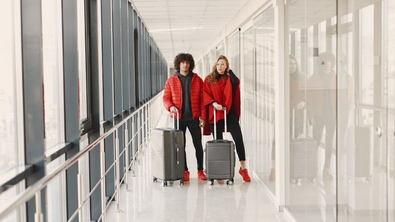 Stylish couple in red outerwear standing in an airport corridor with rolling suitcases.