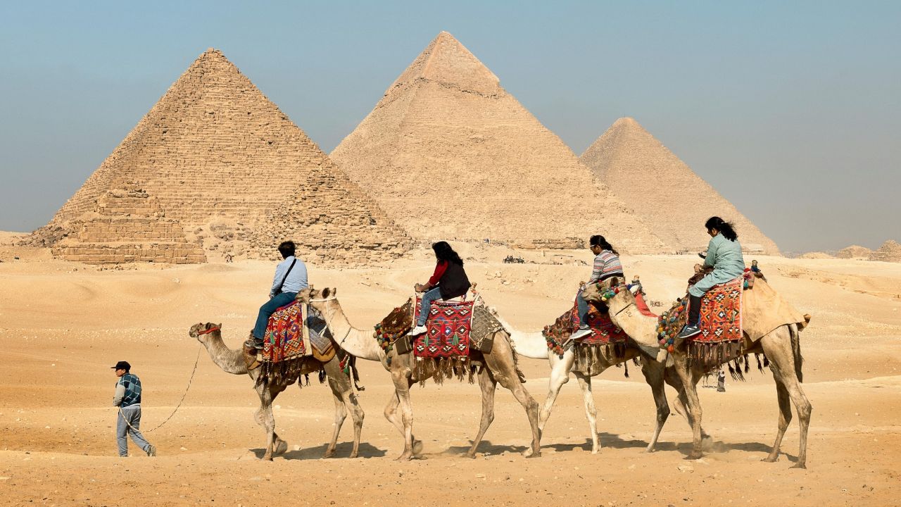 Tourists riding camels near the Pyramids of Giza in Egypt with a guide leading the way.