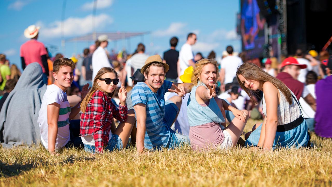 A group of young people sitting on dry grass at an outdoor festival, smiling and enjoying the sunny day.