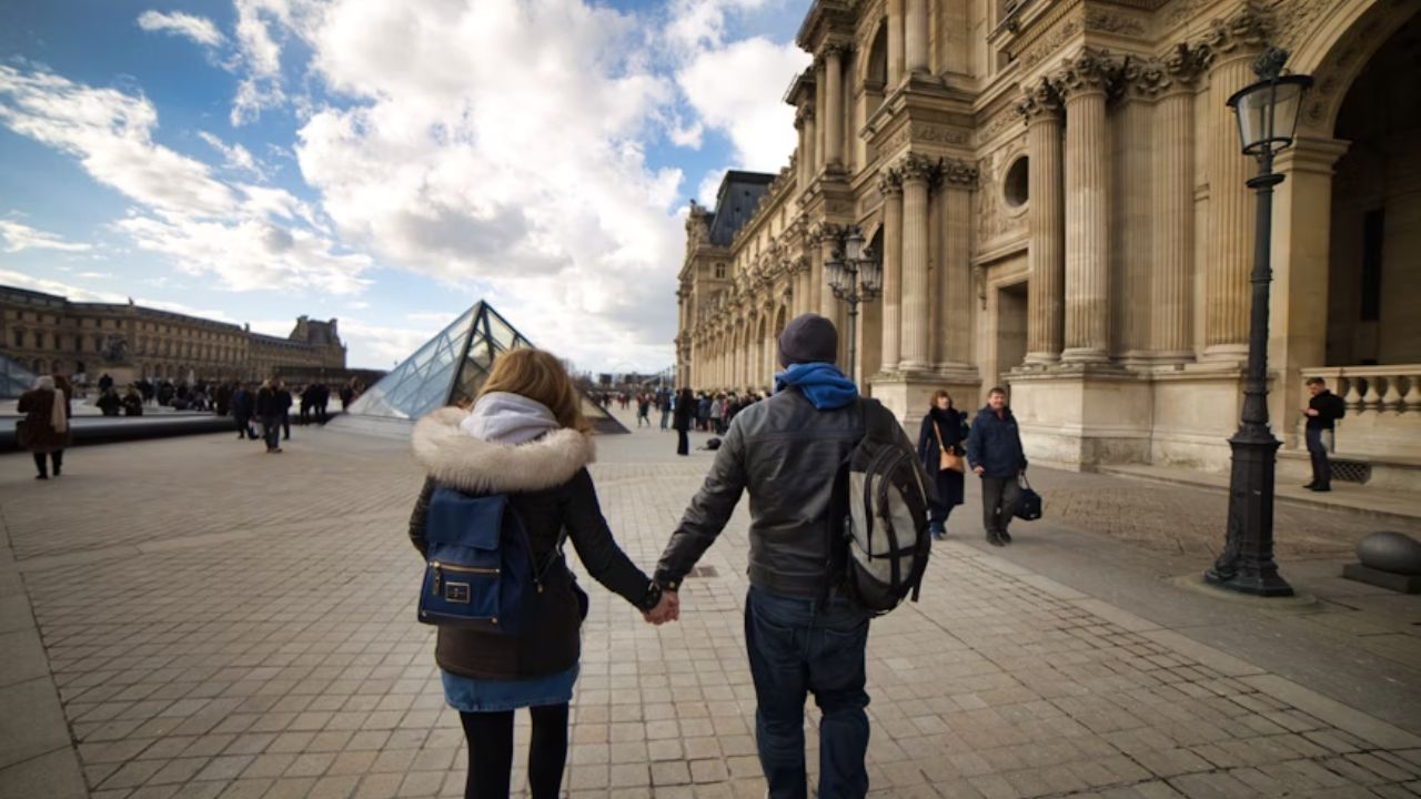 A couple holding hands while walking near the Louvre Museum in Paris, with the glass pyramid and historic architecture in the background.