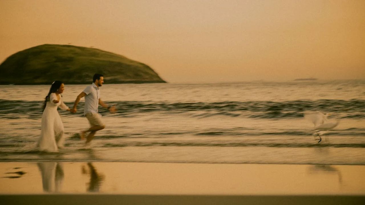 A couple joyfully runs along the beach at sunset, holding hands with waves at their feet and a small island in the background