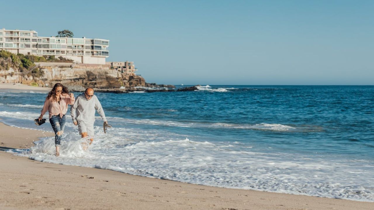 Happy couple walking barefoot along the shoreline at a beach near coastal cliffs and buildings.