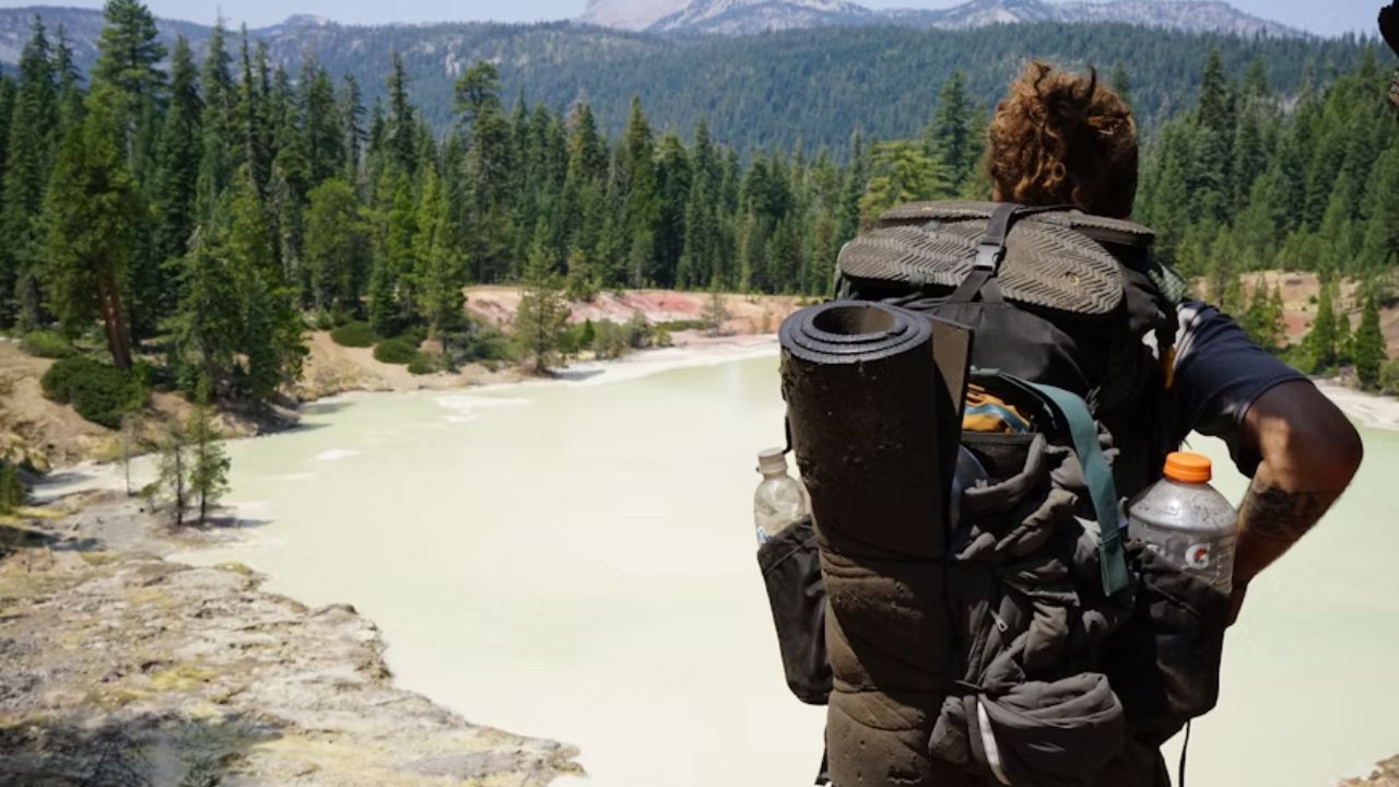 Backpacker with a large hiking pack standing near a geothermal lake surrounded by forest and mountains.