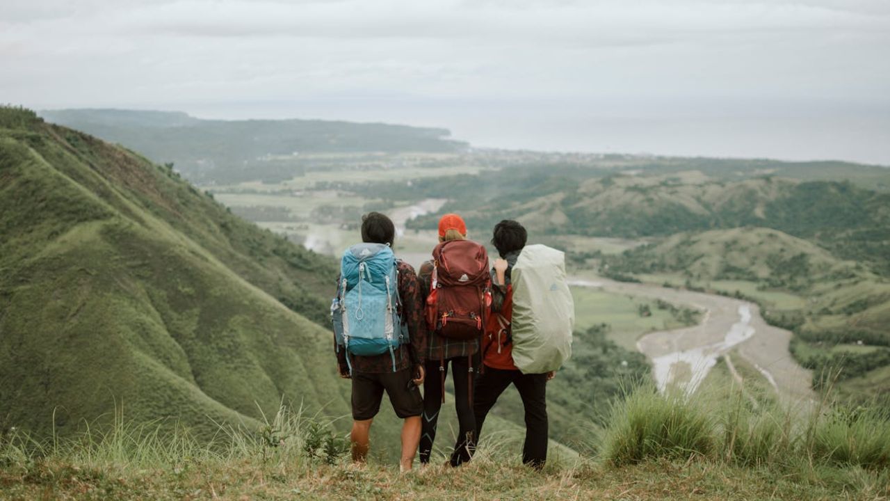 hree hikers with backpacks standing on a grassy hilltop overlooking a scenic valley with rivers and mountains.