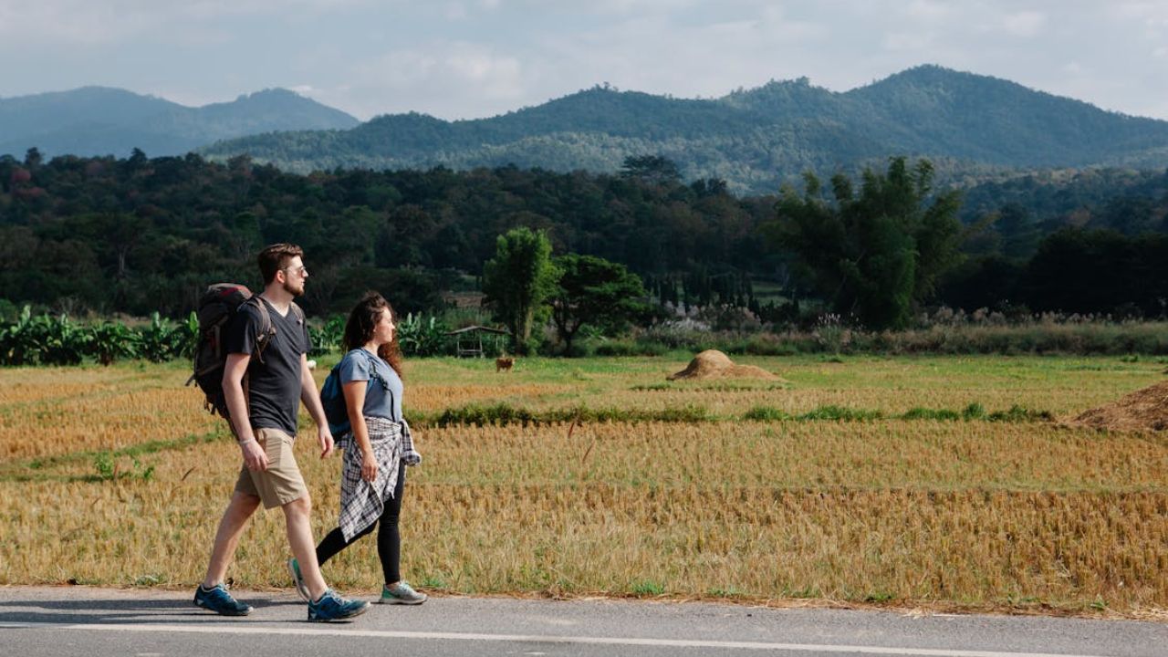Two people walking along a rural road with fields and mountains in the background.