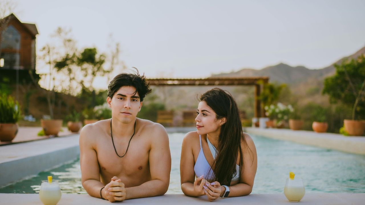 A man and woman sit by an outdoor pool with drinks, mountains, and trees in the background.