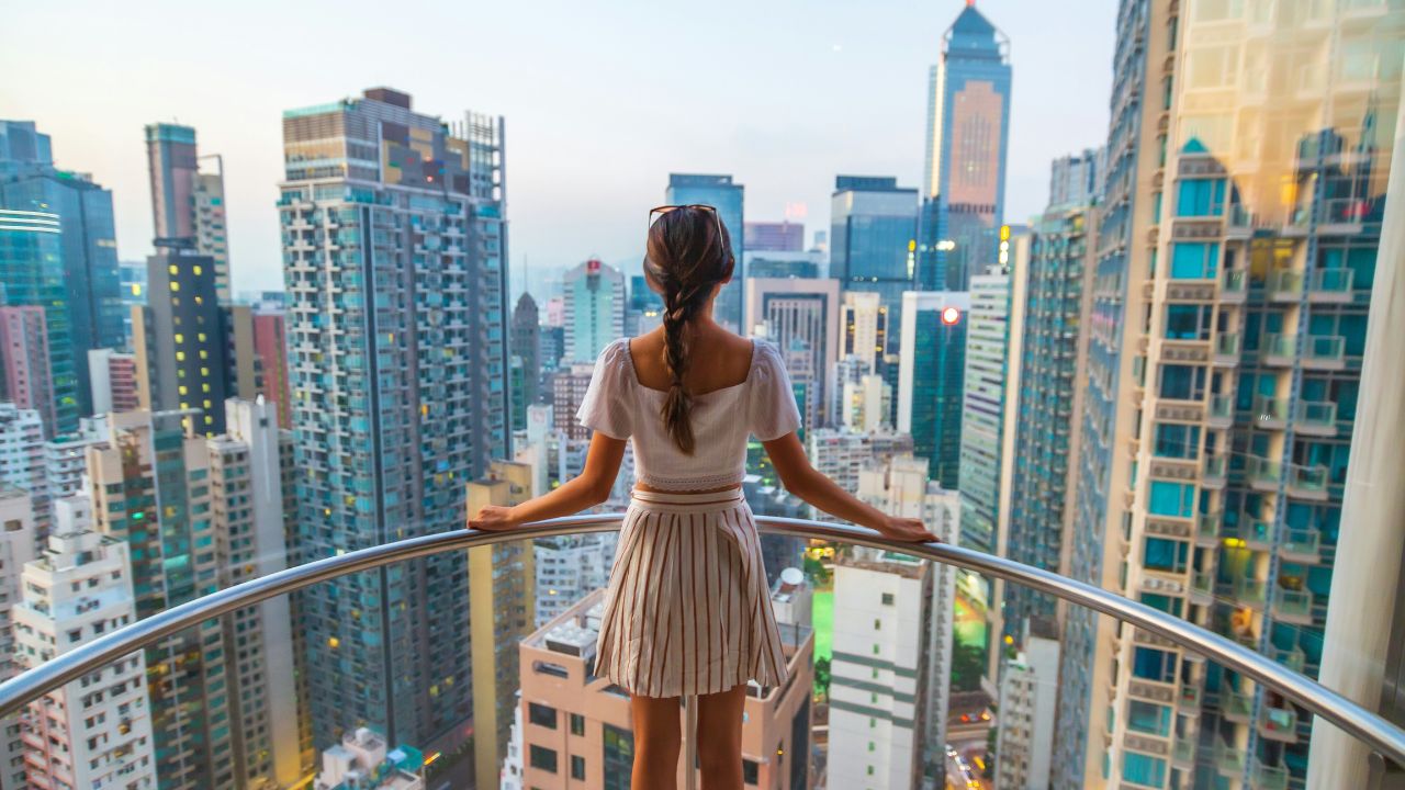 Woman standing on a balcony overlooking a city skyline filled with tall buildings.