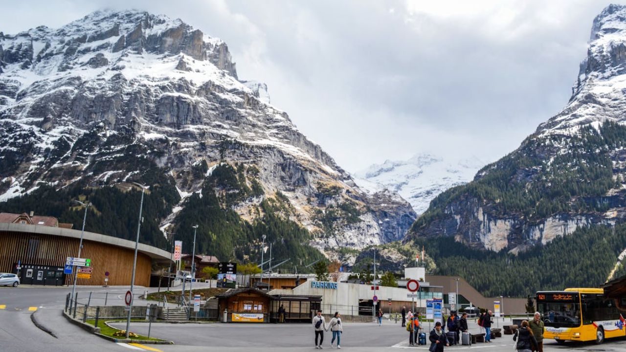 A small mountain village surrounded by snow-covered peaks, with people walking near a bus stop and alpine buildings.