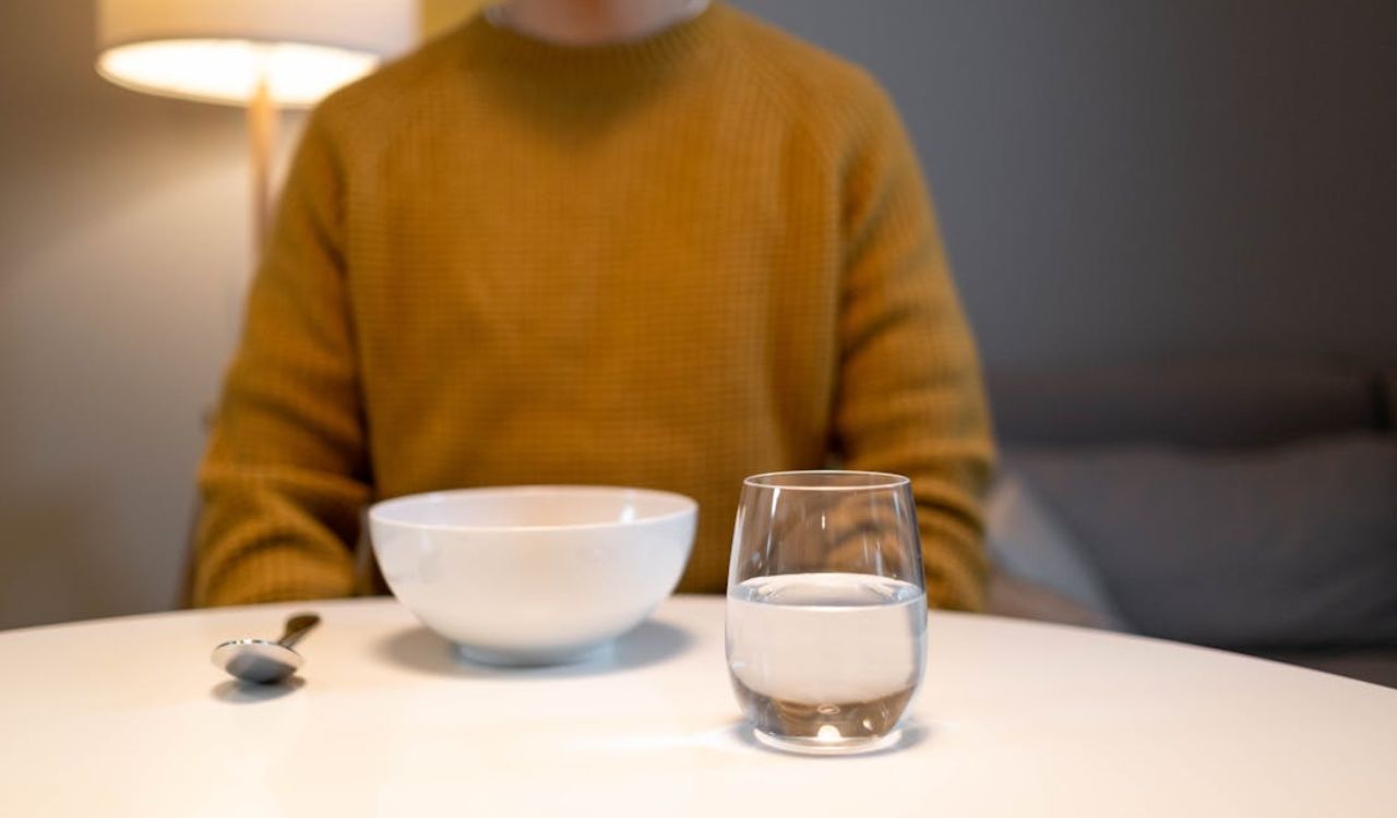 Person in a mustard sweater sitting at a table with a bowl, spoon, and glass of water—highlighting the importance of staying hydrated and eating light