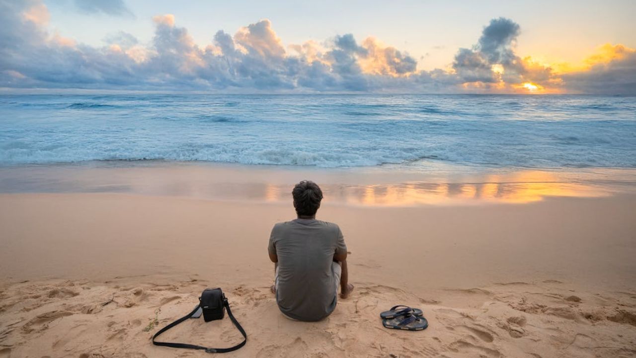 Man sitting alone on a sandy beach at sunset, facing the ocean with a camera bag and sandals beside him.