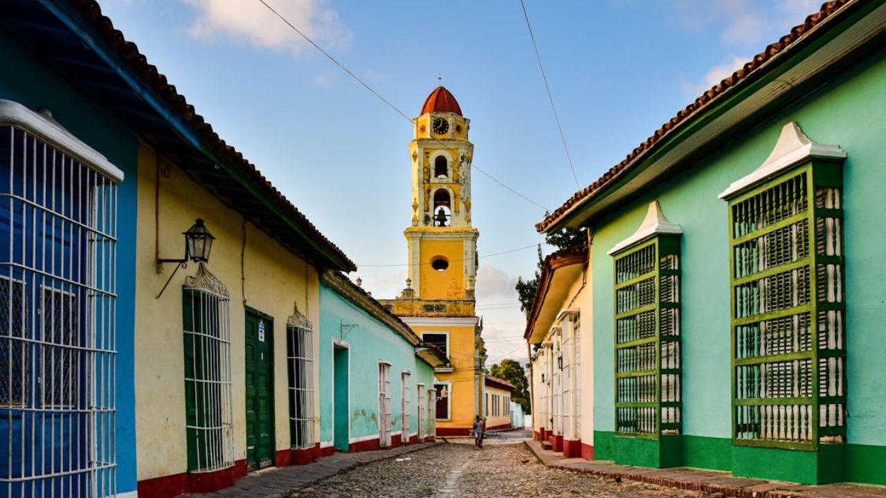 Colorful colonial-style buildings line a cobblestone street leading to a yellow bell tower under a blue sky in Trinidad, Cuba.