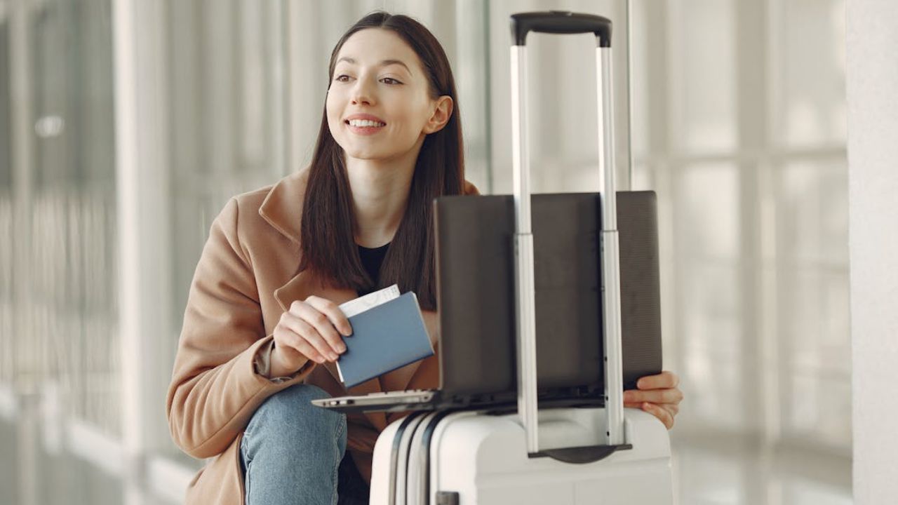 A smiling woman at an airport holding a passport and boarding pass, sitting beside a suitcase with a laptop on top.