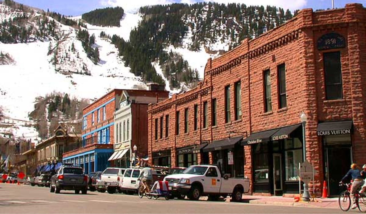Historic red-brick buildings and parked cars line a street in Aspen, Colorado, with snow-covered ski slopes in the background
