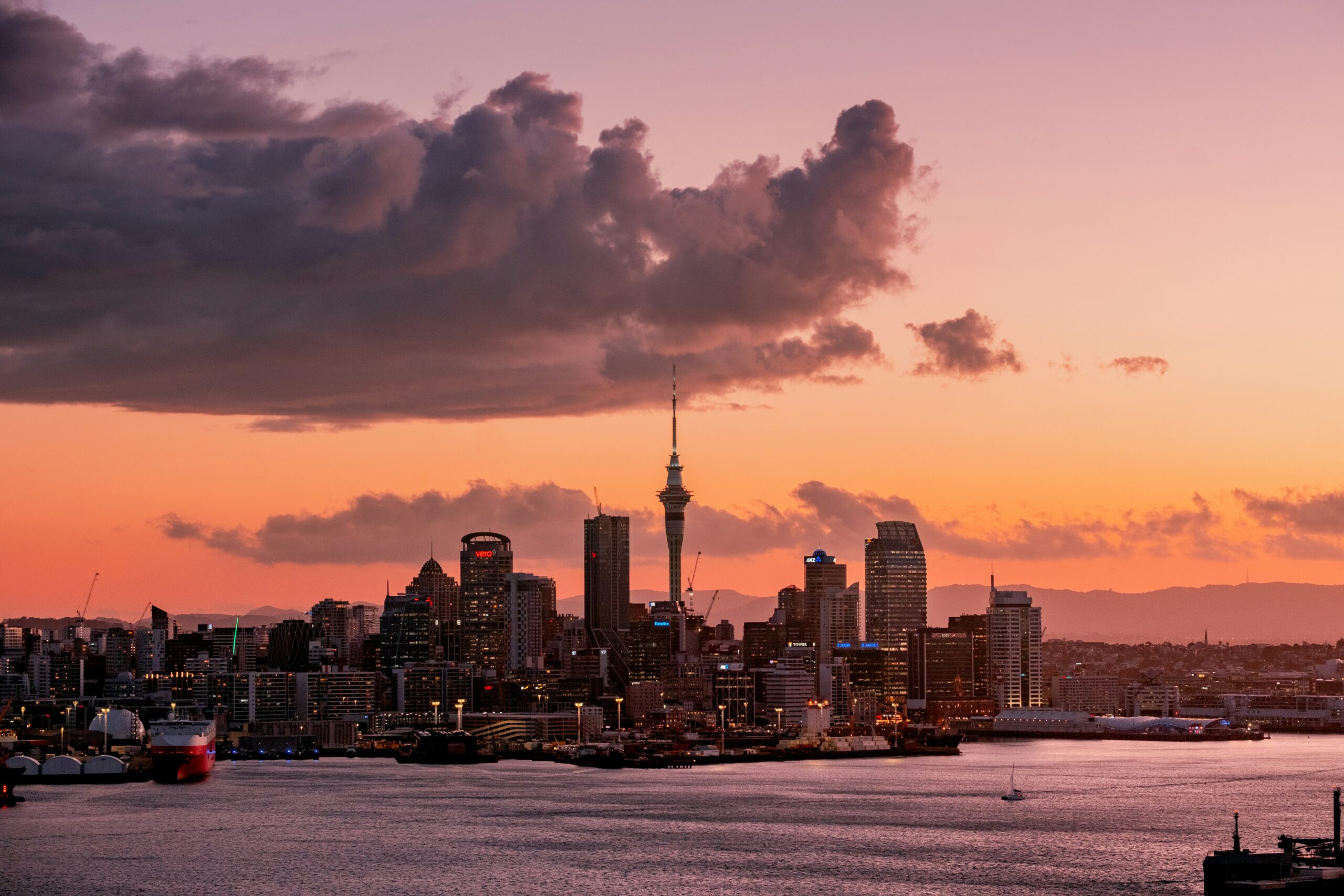 A city skyline with tall buildings and a prominent tower is seen across the water at sunset beneath a large cloud.